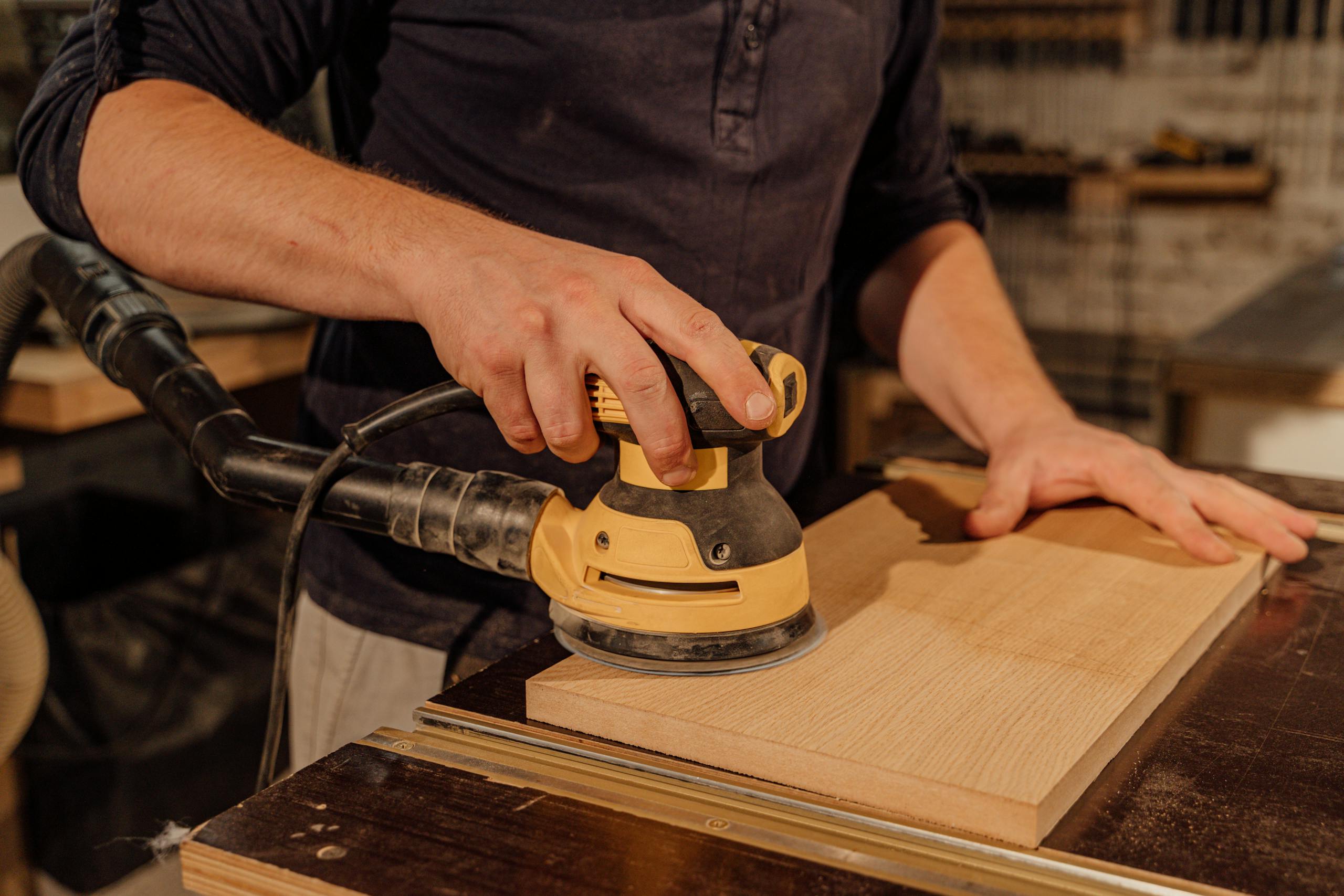 Carpenter using an electric sander on a wooden board in a workshop, showcasing craftsmanship.
