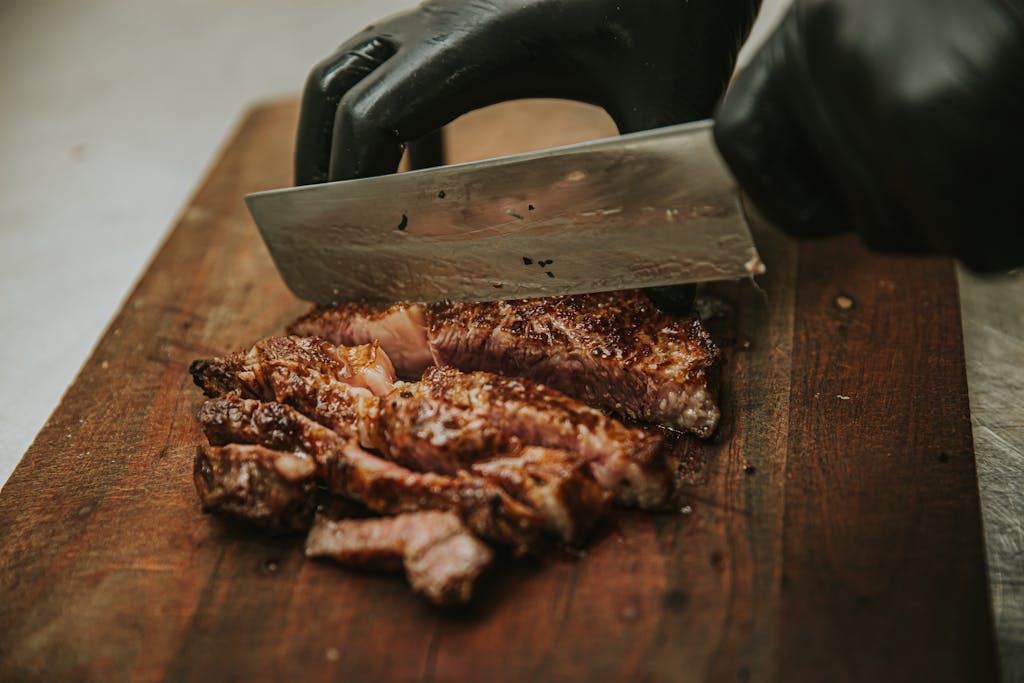 Close-up of chef slicing juicy grilled steak on wooden cutting board with cleaver.