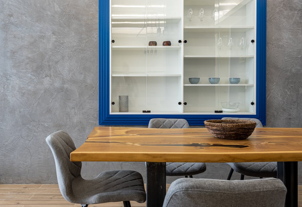 Comfortable chairs at wooden dinner table with bowl placed near gray wall with glass cabinet with glassware and dishes in modern kitchen