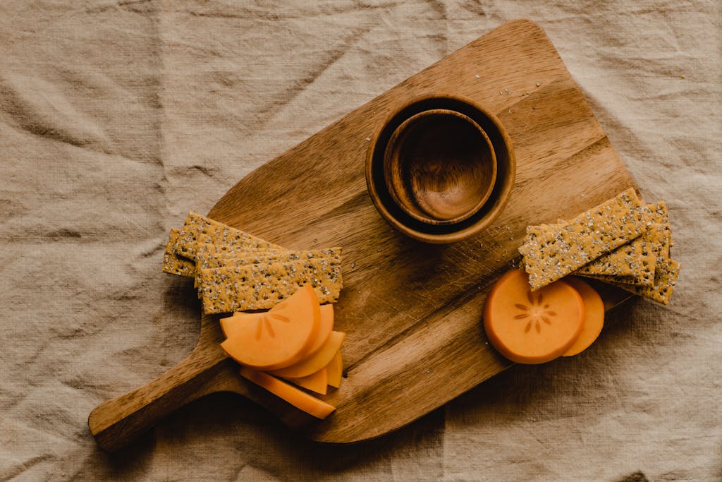 Flat lay of wood board with sliced persimmon, crackers, and bowls for a rustic vibe.