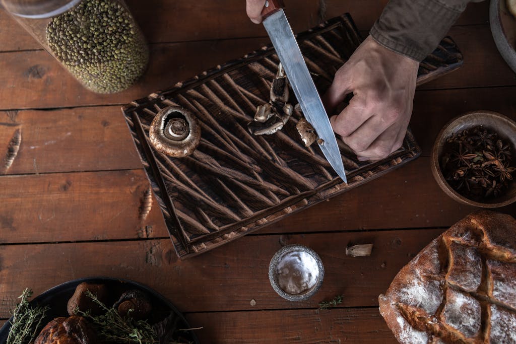 Top-down view of hands slicing mushrooms on a cutting board. Rustic wood setting.
