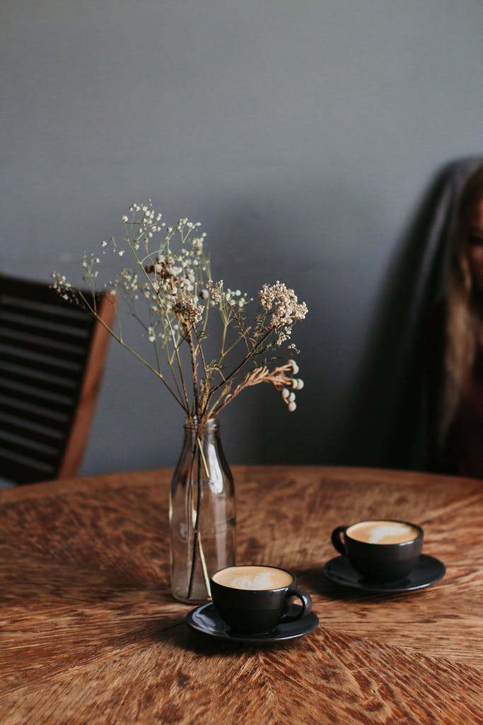 Two cappuccino cups with frothy art on a wooden table, accompanied by a delicate flower vase.